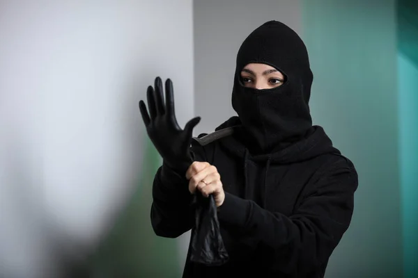 Thief broke into the apartment. House robbery by woman in a black jacket and black mask black gun and crowbar. Burglar in a mask. Thief in a mask trying to break into other people's house.