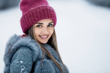 Gri kürklü ve kırmızı şapkalı tatlı bir kızın portresi. Karlı bir günde kış ormanında yürüyor. Çam ormanında kış havası. Noel zamanı açık hava.