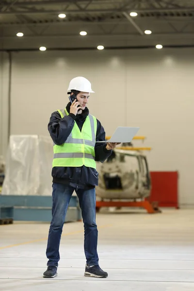 Young man, factory engineer in a work dress and white hard hat holding smartphone and laptop organizing and controlling work process in a industrial factory.