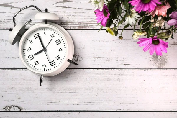 Beyaz ahşap zemin üzerinde çiçekler olan beyaz alarmlı saatin üst görüntüsü. Bahar geldi..