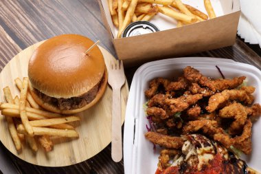 Plastik kaplarda yiyecek dolu bir masa. Yemek servisi. Tavuk salatası, soslu tavuk kanadı, ahşap masada patates kızartması üzerine hamburger. İş yemeği..