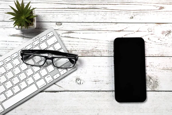 Flat lay view of white keyboard, smartphone and black glasses on a white wooden background. Top view of modern office workplace.