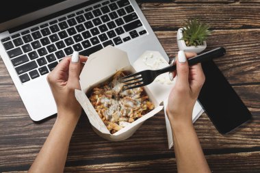 Kadın elleri dizüstü bilgisayarın yanındaki tahta masada yemek kabını açıyor. Fast food teslimatı. Evde iş yemeği yiyorum. Evden çalışıyorum. Uzak çalışma