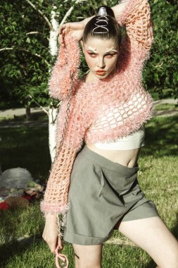 Örgü giyip güneş gözlüğü takan manken yazın dışarıda poz veriyor. Yaratıcı saç stili ve profesyonel makyajlı güzel genç bir kadın pembe aksanlı canlı doğal çevreden hoşlanıyor. Yumuşak doğal aydınlatma vurguluyor