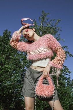 Örgü giyip güneş gözlüğü takan manken yazın dışarıda poz veriyor. Yaratıcı saç stili ve profesyonel makyajlı güzel genç bir kadın pembe aksanlı canlı doğal çevreden hoşlanıyor. Yumuşak doğal aydınlatma vurguluyor