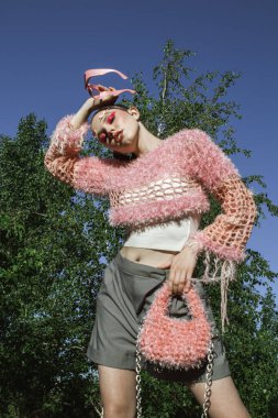 Örgü giyip güneş gözlüğü takan manken yazın dışarıda poz veriyor. Yaratıcı saç stili ve profesyonel makyajlı güzel genç bir kadın pembe aksanlı canlı doğal çevreden hoşlanıyor. Yumuşak doğal aydınlatma vurguluyor