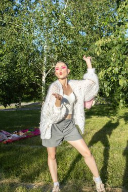 Yazın dışarıda poz veren beyaz örgü kazaklı güzel bir manken. Yaratıcı saç stili ve profesyonel makyajlı şık genç bayan aydınlık bir yaz gününün tadını çıkarıyor. Yumuşak doğal ışıklandırma örgü giysisinin fotorealistik dokusunu vurgular