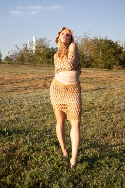 Yazın bej örgü bluz ve etek giyen manken. Profesyonel makyajlı genç bir kadın elinde el yapımı bir kova şapkayla dışarıda poz veriyor. Yumuşak doğal ışık örgü ve derinin gerçekçi dokusunu vurgular.