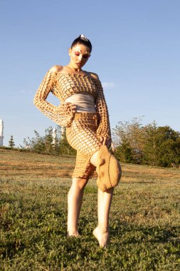 Yazın bej örgü bluz ve etek giyen manken. Profesyonel makyajlı genç bir kadın elinde el yapımı bir kova şapkayla dışarıda poz veriyor. Yumuşak doğal ışık örgü ve derinin gerçekçi dokusunu vurgular.