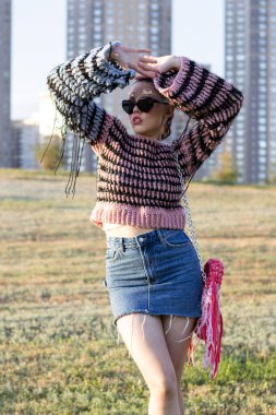 Yazın örgü süveter ve püsküllü bluz giyen manken. Etekli ve güneş gözlüklü şık genç bir kadın, elinde el yapımı bir çantayla dışarıda poz veriyor. Yaratıcı yaz fotoshoot el yapımı dokuları, moda aksesuarları ön plana çıkarıyor