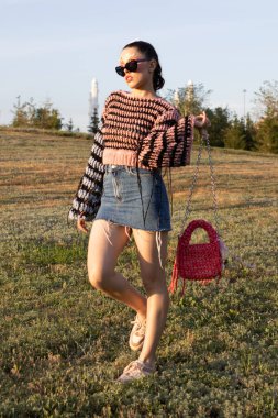 Yazın örgü süveter ve püsküllü bluz giyen manken. Etekli ve güneş gözlüklü şık genç bir kadın, elinde el yapımı bir çantayla dışarıda poz veriyor. Yaratıcı yaz fotoshoot el yapımı dokuları, moda aksesuarları ön plana çıkarıyor