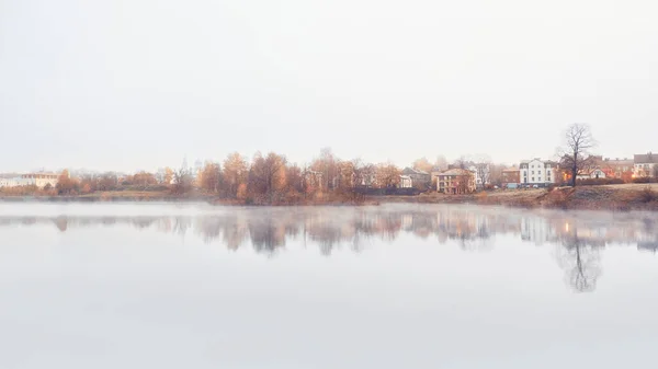 Panorama mglistego porannego minimalistycznego krajobrazu z linią domków i drzew nad wodą jesienią. — Zdjęcie stockowe
