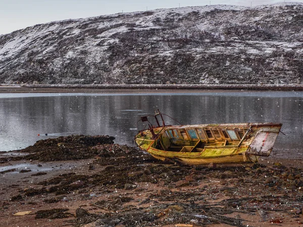 An old rusty fishing boat washed up on a sandy beach in the Barents Sea. Authentic the North sea. Pollution of the coastline. Russia.