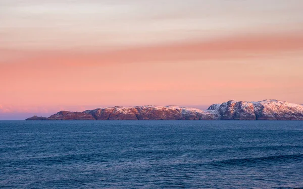 Beautiful pink sunset on the Arctic Ocean. A peninsula with snow-capped hills on the horizon. Sea minimalistic landscape. Panoramic view.