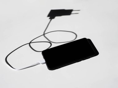 Beyaz bir masada gölgeli şarj kablosu olan siyah bir akıllı telefon. Üst görünüm.