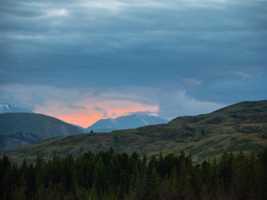 Dağlarda şafak vakti. Gün batımlı ya da gündoğumlu renkli doğa manzarası. Atmosferik manzara dağların siluetleriyle portakal rengi gökyüzünün arka planında ağaçlar. 
