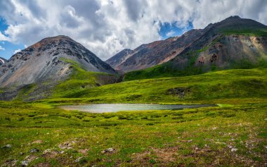 Dağlık yeşil vadideki dereleri bulutlu gökyüzünün altındaki daha büyük dağlardan gelen güzel sığ dağ gölü ile panoramik alp manzarası. Aynalı buzul gölü ile yağmurlu dağ manzarası.