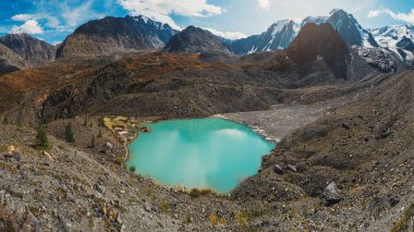 Dağlık vadide, güneş ışığında dağ gölü ve mavi bulutlu gökyüzünün altında büyük bir dağ manzarası. Sonbahar vadisinde bulutların gölgesi. Panoramik görünümler.