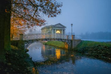 Gece parkının mistik manzarası, nehir kanalı üzerinde antik bir köprü ve penceresinde ışık olan bir pavyon, gece sisi. Gatchina. Rusya.