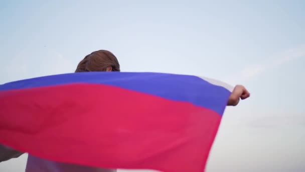 Das Gesicht einer schönen Frau mit einer russischen Flagge in den Händen. Hübsche Frau läuft mit Fahne auf dem Rücken gegen den Sonnenuntergang — Stockvideo