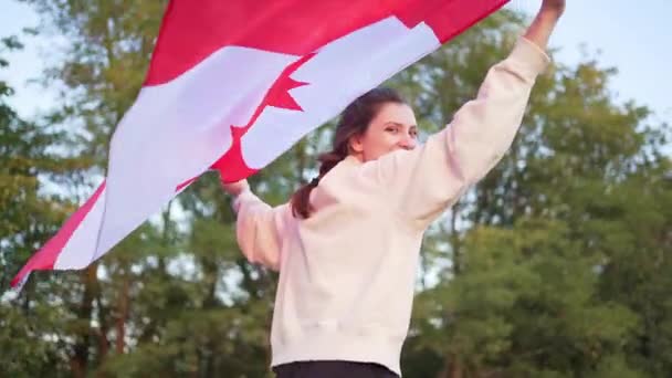 一个拿着加拿大国旗的年轻貌美的女子与森林的背景正好相反。加拿大爱国者笑着看着摄像机 — 图库视频影像