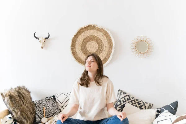 Un estilo de vida saludable. Una mujer descansa con ropa casual en una habitación auténtica. Postura de loto en el sofá, meditación —  Fotos de Stock