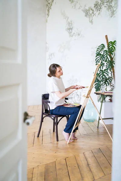 Pintor en un taller. Vista desde la puerta. Joven artista femenina dibuja sentado frente al caballete —  Fotos de Stock