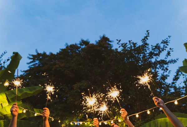 Patio party. The hands of different people hold sparklers. New Year in the tropics. Friends dancing and having fun