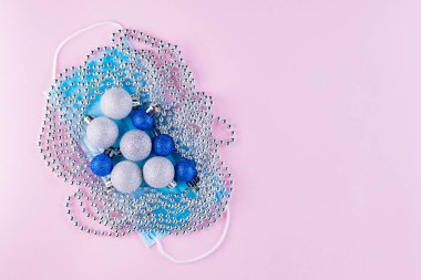 Yüz maskesinde noel boncukları ve toplar. Pembe arka planda Noel ağacı süslemeleri olan tıbbi maske. Coronavirus Noel geçmişi. Uzayı kopyala Düz yatıyordu