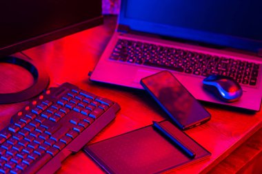 Dijital tasarımcı işyeri. Uzaktan kumanda. Neon ışığı altında serbest çalışan bir işyeri. Bilgisayar, dizüstü bilgisayar, grafik tableti ve akıllı telefon masada.