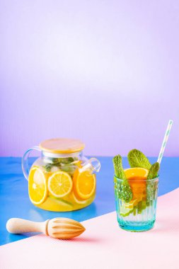 Limonata, limon, portakal ve nane yapraklarından oluşan çok renkli bir arka plan. Şeffaf çaydanlıkta limonlu buzlu çay. Yaratıcı bileşim