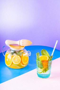 Limonata, limon, portakal ve nane yapraklarından oluşan çok renkli bir arka plan. Şeffaf çaydanlıkta limonlu buzlu çay. Yaratıcı bileşim