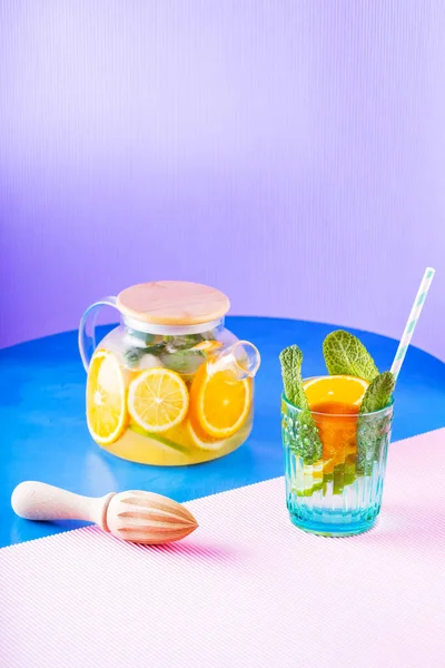 Limonata, limon, portakal ve nane yapraklarından oluşan çok renkli bir arka plan. Şeffaf çaydanlıkta limonlu buzlu çay. Yaratıcı bileşim