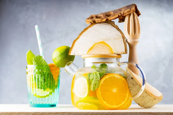 Lemonade drink of water, lemon, orange and mint leaves in a transparent teapot. Lime mint iced tea and pieces of wood. Creative composition