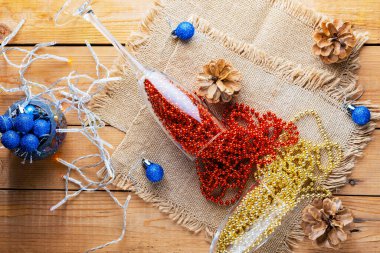 Noel boncuklu iki şampanya bardağı. Noel baloları ve çuval bezinin üzerindeki süslemeler. Ahşap arka planda çam kozalakları ve çok renkli boncuklar. Üst görünüm