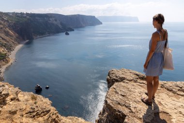 Genç bir kadın kayanın tepesinde duruyor ve yazın renkli günbatımında deniz kıyısına ve dağlara bakıyor. Kız, deniz, dağ tepeleri ve güneşli turuncu gökyüzü manzarası. Oludeniz, Türkiye