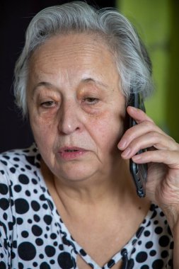 Yaşlı, beyaz saçlı, emekli bir kadın cep telefonuyla konuşuyor. Evdeki masada oturuyor.