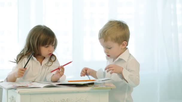 Les petits enfants jouent avec des crayons. Garçon et fille dessinent près de la fenêtre. Enfants en vêtements brillants tableaux peints dans les livres sur la table . — Video