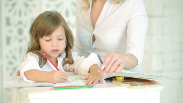 Little girl drawing at the table with her mother. A child dressed in white paints pencils drawing. On the background of the window and white curtains. Carvings of the wall. European mom and baby girl.