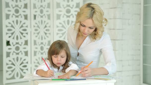 Маленькая девочка рисует за столом со своей матерью. Ребенок, одетый в белые краски, рисует карандаши. На заднем плане окна и белые занавески. Резьба по стене. Европейская мама и девочка . — стоковое видео