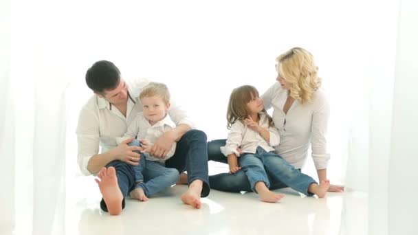 Feliz família pai mãe menina e menino sentado no chão. A abraçar-nos. Pai com filho, mãe com filha, a sorrir, a olhar um para o outro. Cortinas de luz e a janela. Muita luz. . — Vídeo de Stock