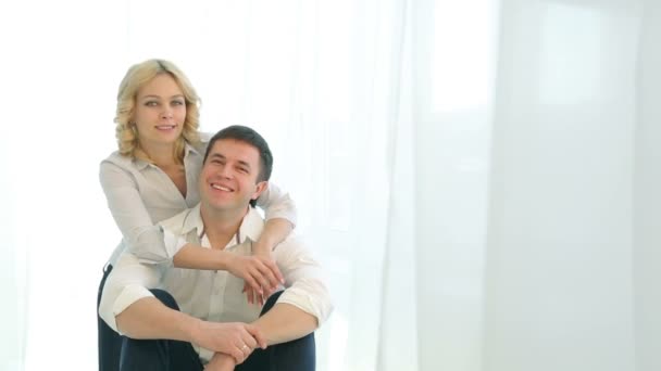 大きな窓の近くで隣同士の床に座る夫婦は、カメラを見て抱き合う。明るいカーテンが備わります。白いシャツとジーンズで幸せな家族。笑顔の男女. — ストック動画