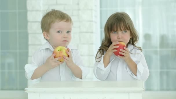 El niño y la niña comen manzanas rojas. Nos miramos el uno al otro y luego nos salimos de cuadro. Niños en camisas blancas al lado de la mesa en una habitación luminosa. Sobre el fondo de la pared de ladrillo claro y la ventana . — Vídeo de stock