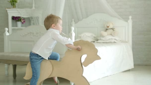 Un niño montado en un caballo de madera. Chico sonríe. El niño en el dormitorio contra una cama blanca con dosel. Sobre el fondo de la pared de ladrillo blanco y una cómoda. Dormitorio . — Vídeos de Stock