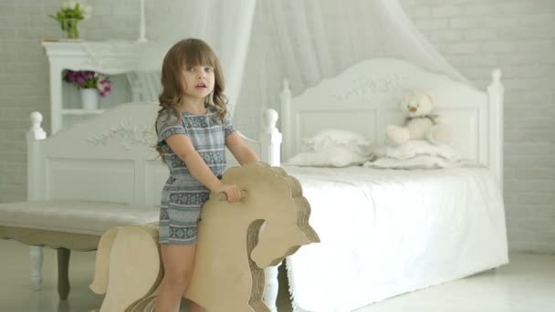 Una niña montado en un caballo de madera. La chica sonríe. La niña en el dormitorio contra una cama blanca con dosel. Sobre el fondo de la pared de ladrillo blanco y una cómoda. Dormitorio . — Vídeos de Stock