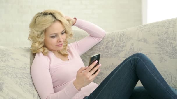 Schönes Mädchen mit Smartphone auf dem Sofa liegend. helles europäisches Mädchen in rosa Pullover. auf dem Hintergrund von weißer Ziegelwand und Fenster. — Stockvideo