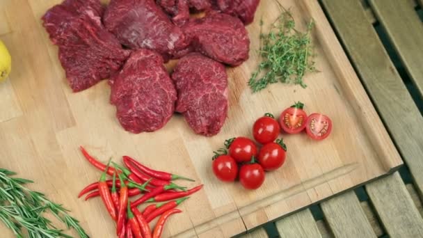 Carne fresca para filete sobre tabla de madera. Carne de mármol, carne de res. Cerca de tomates, romero, tomillo, limón, pimiento rojo, arándano. El cocinero vierte el arándano en el tablero, añade romero a la carne . — Vídeo de stock