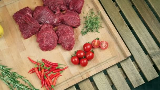 Viande fraîche pour steak sur planche de bois. Viande marbrée, bœuf. Près de tomates, romarin, thym, citron, poivron rouge, canneberge. Le cuisinier verse la canneberge sur le plateau, ajoute du romarin à la viande . — Video