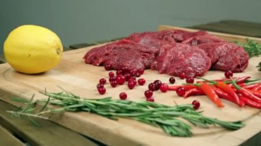 Ahşap tahta üzerinde biftek için taze et. Mermer et, sığır eti. Domates, biberiye, kekik, limon, kırmızı biber, kızılcık. Aşçı kızılcık tahtada dökülen, biberiye ete ekler.