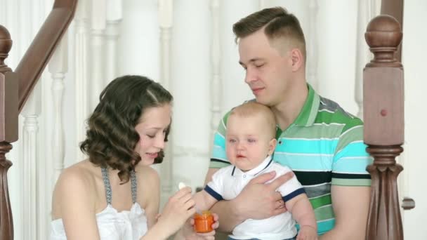 De moeder voedt het kind en vader houdt hem. Gelukkige familie. Mama papa en 6 maanden oude baby. Familie zittend op de trap thuis. — Stockvideo